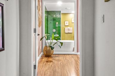 Hallway view into a modern bathroom featuring a freestanding white tub behind glass, glossy green tile shower wall, olive-yellow accent wall with framed art, wood-look flooring and a potted plant in a wicker basket.
