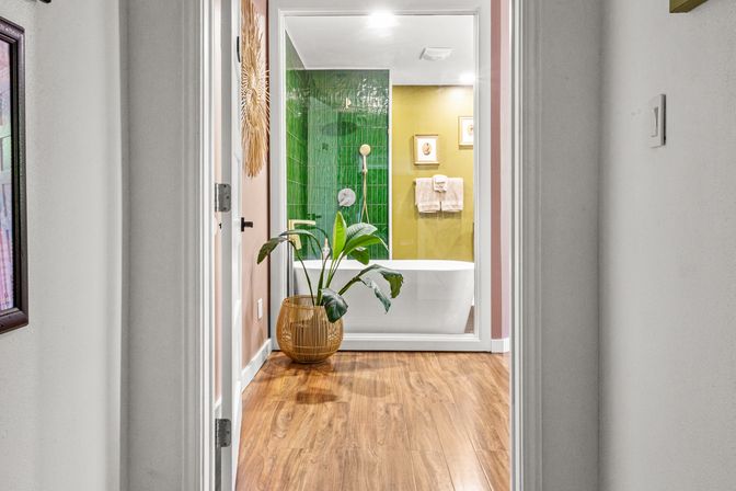 Hallway view into a modern bathroom featuring a freestanding white tub behind glass, glossy green tile shower wall, olive-yellow accent wall with framed art, wood-look flooring and a potted plant in a wicker basket.