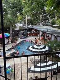 Overhead view of a lively outdoor pool party with swimmers and loungers on a tiled patio, black-and-white striped umbrellas, a colorful beach umbrella, cabana seating, palm trees and dozens of floating soap bubbles.
