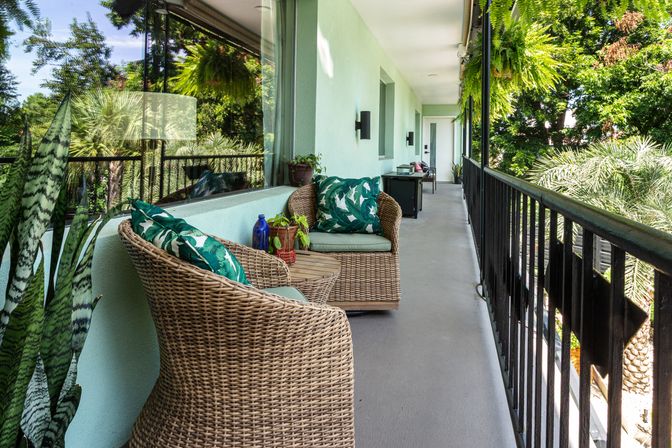 Cozy sunny balcony with wicker armchairs and green leaf-pattern cushions, small round table with potted plants and decorative bottles, black metal railing and hanging ferns overlooking lush palm trees.