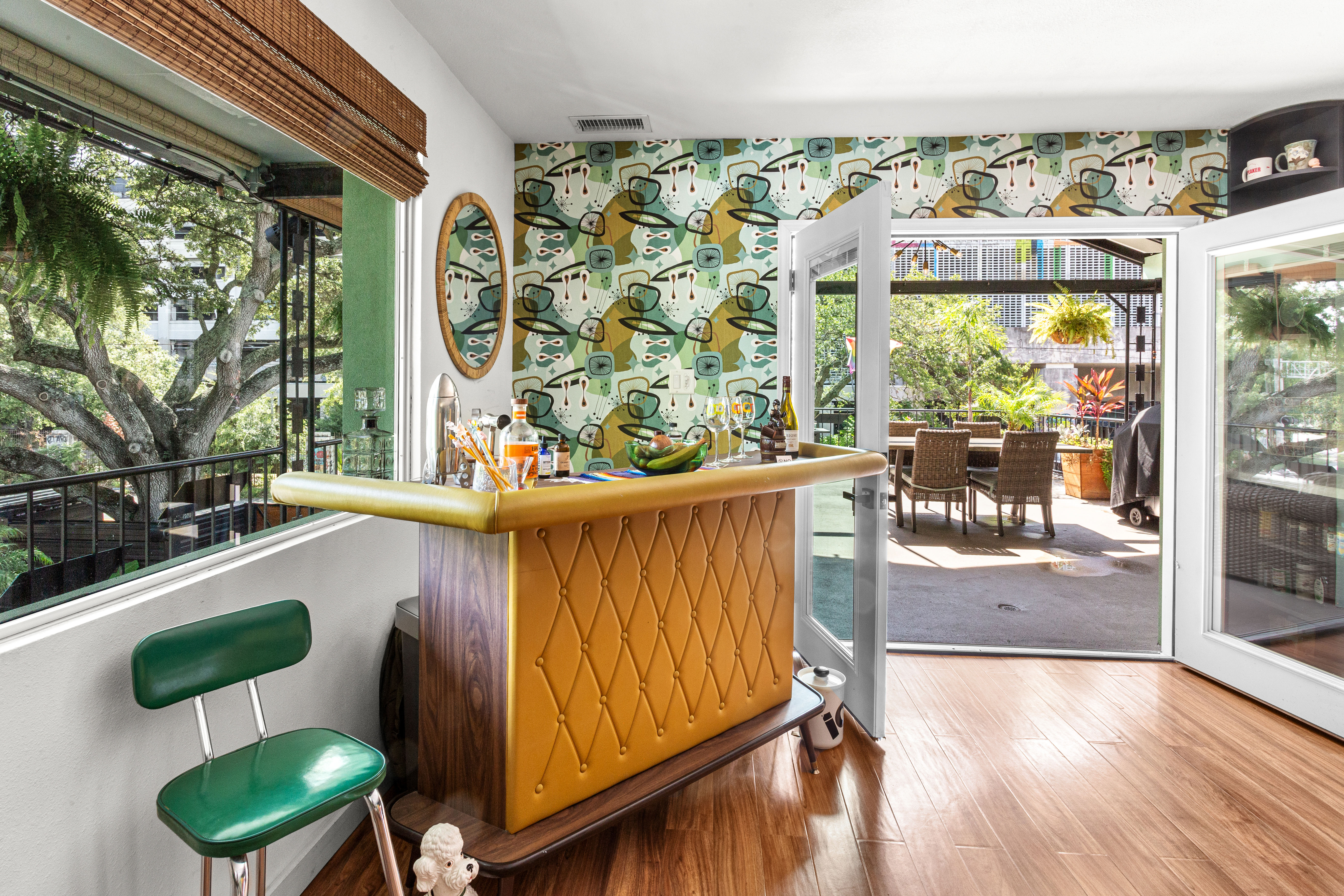 Sunlit retro home bar with tufted mustard front, green vinyl stool and mid-century abstract wallpaper, opening through glass doors to a leafy patio dining area under large trees.