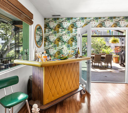Sunlit retro home bar with tufted mustard front, green vinyl stool and mid-century abstract wallpaper, opening through glass doors to a leafy patio dining area under large trees.