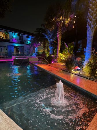 Nighttime tropical hotel pool courtyard with palm trees and lounge chairs lit by vibrant neon blue, purple, and orange LED lights, dancing fountain jets and colorful reflections on the water