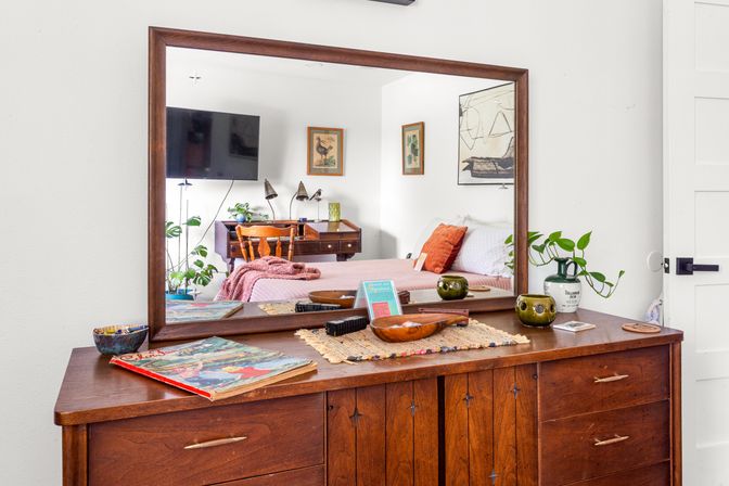 Mid-century wooden dresser with a large mirror reflecting a cozy bedroom—pink bedspread, wooden desk, wall art and TV—dresser top styled with potted plants, ceramic bowls, books and a woven runner