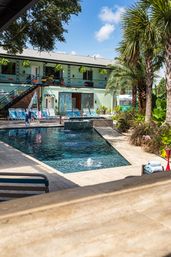 Sunny tropical hotel courtyard with a rectangular outdoor pool, bubbling fountain, palm trees, striped lounge chairs and a mint-green two-story building with hanging plants.