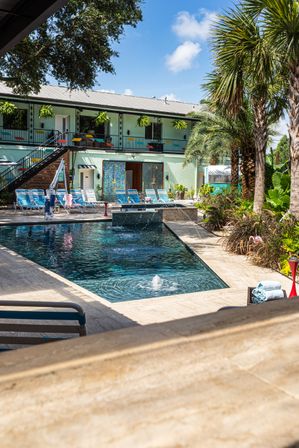 Sunny tropical hotel courtyard with a rectangular outdoor pool, bubbling fountain, palm trees, striped lounge chairs and a mint-green two-story building with hanging plants.