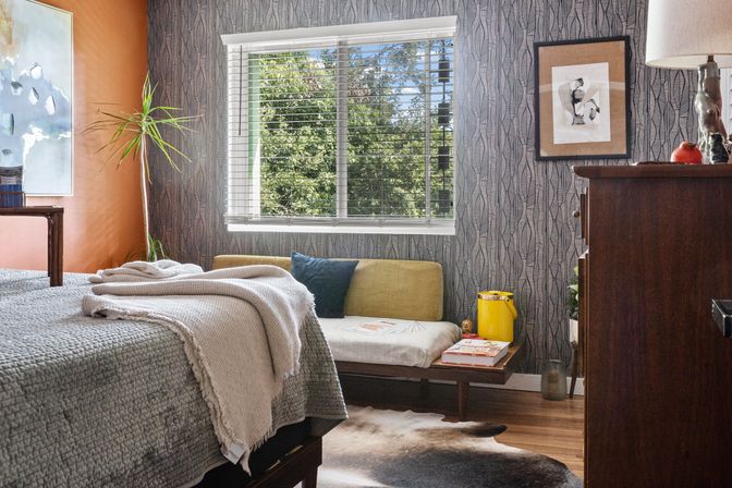 Sunlit mid-century modern bedroom interior with patterned wallpaper and orange accent wall, window with white blinds overlooking leafy trees, textured gray bedding with creamy throw, low daybed with yellow cushion, wooden dresser, potted plant and cowhide rug.