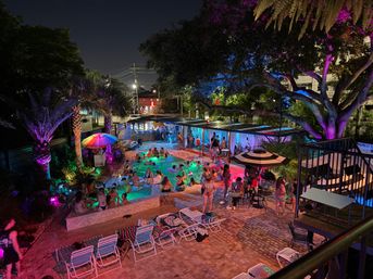 Nighttime outdoor pool party with colorful LED uplighting, palm trees and a large illuminated tree, swimmers and groups socializing around cabanas, umbrellas and lounge chairs on a brick patio — urban poolside nightlife scene.
