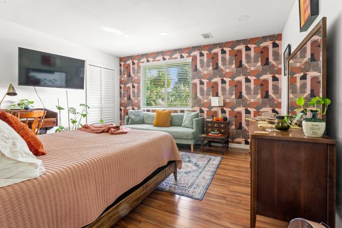 Sunny mid-century modern bedroom with bold geometric accent wall, hardwood floors, dusty-rose quilted bed, green velvet loveseat under a window with blinds, wooden dresser, plants, area rug and wall-mounted TV.