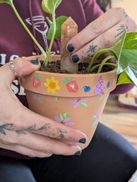 Hand-painted terracotta pot decorated with flowers and strawberries holding a pothos plant and a quartz point crystal, held by tattooed hands with black nail polish — DIY indoor plant decor
