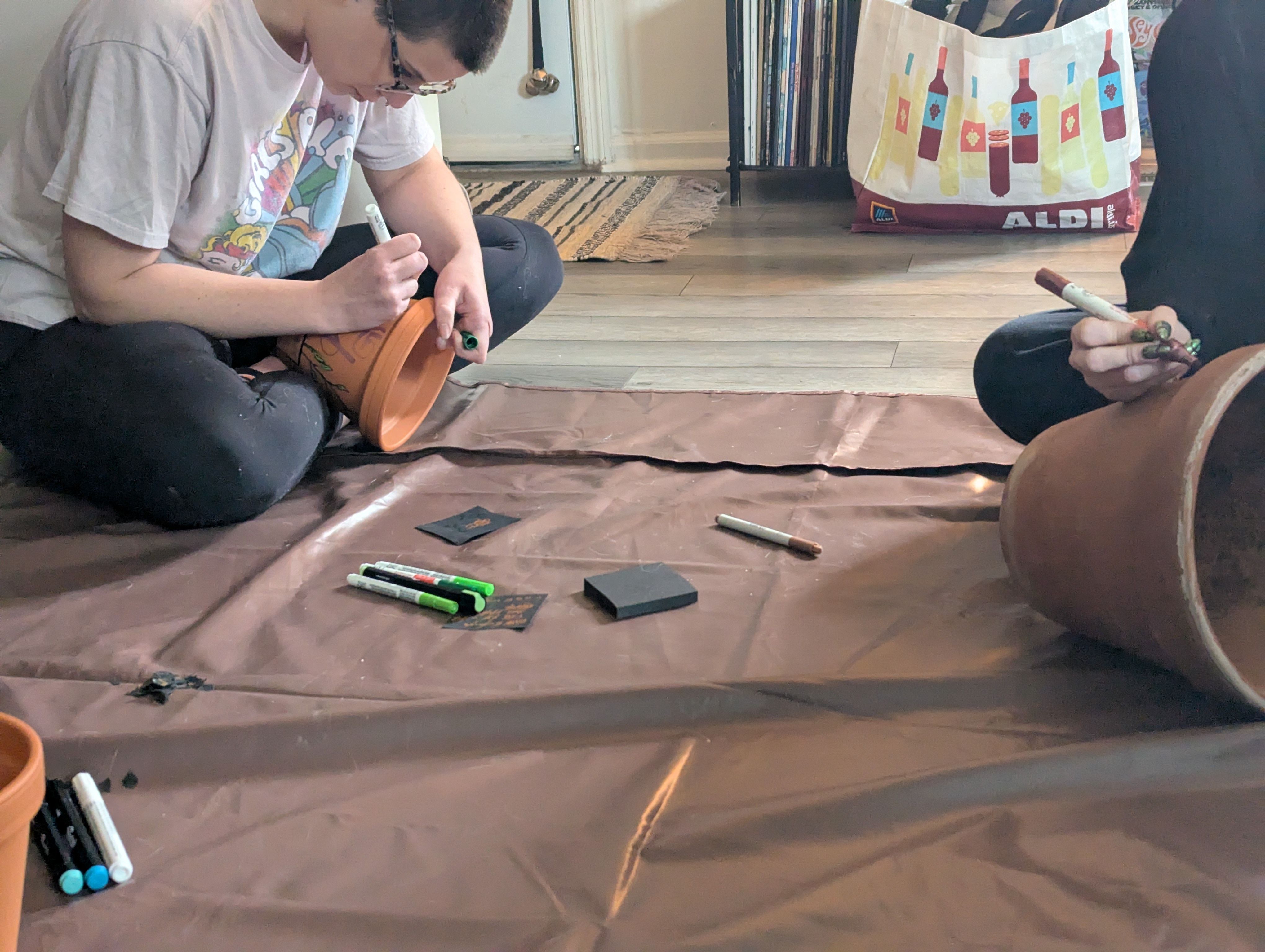 Two people sitting on a floor painting terracotta plant pots with markers and art supplies on a brown drop cloth in a cozy indoor DIY crafting session.