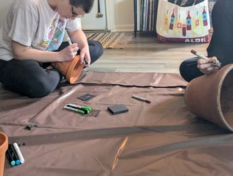 Two people sitting on a floor painting terracotta plant pots with markers and art supplies on a brown drop cloth in a cozy indoor DIY crafting session.
