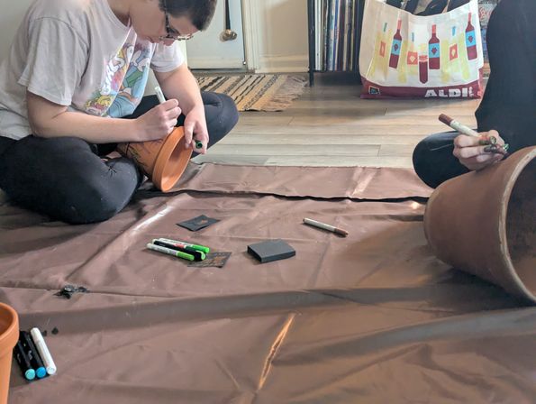 Two people sitting on a floor painting terracotta plant pots with markers and art supplies on a brown drop cloth in a cozy indoor DIY crafting session.