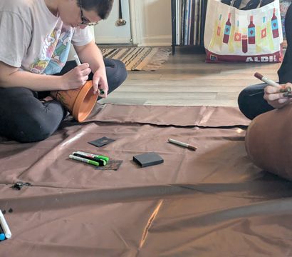 Two people sitting on a floor painting terracotta plant pots with markers and art supplies on a brown drop cloth in a cozy indoor DIY crafting session.