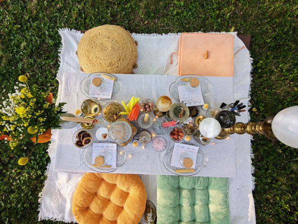 Overhead view of a boho garden picnic: low white table on grass with candles, jars, dried citrus, fresh yellow flowers and colorful tufted floor cushions.