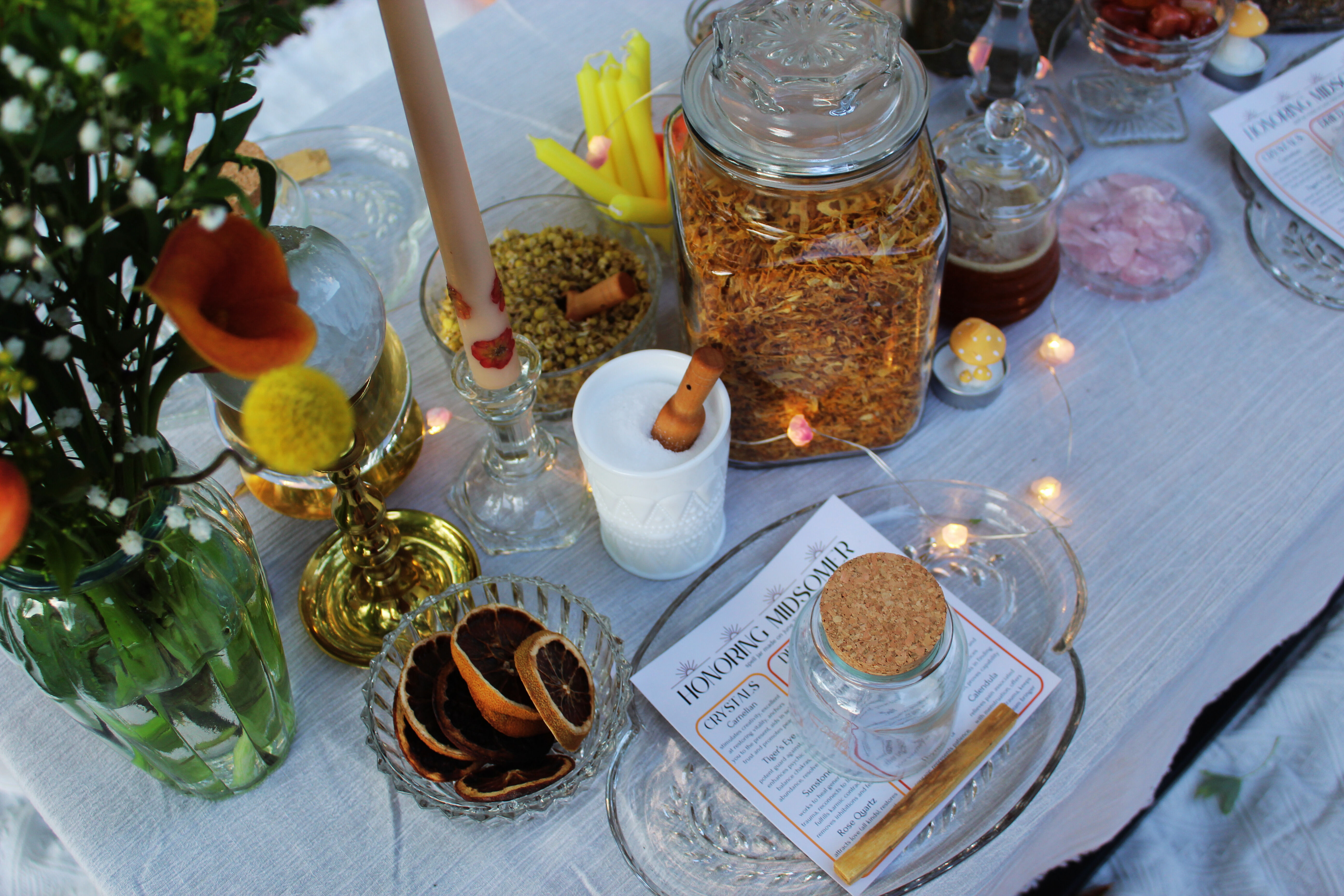 Festive tablescape with glass jars of dried herbs and spices, a corked jar and bowl of dried citrus slices, lit candlesticks, wildflowers and fairy lights on a linen table.