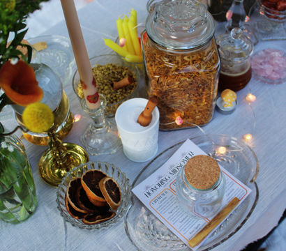 Festive tablescape with glass jars of dried herbs and spices, a corked jar and bowl of dried citrus slices, lit candlesticks, wildflowers and fairy lights on a linen table.