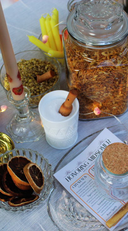 Festive tablescape with glass jars of dried herbs and spices, a corked jar and bowl of dried citrus slices, lit candlesticks, wildflowers and fairy lights on a linen table.