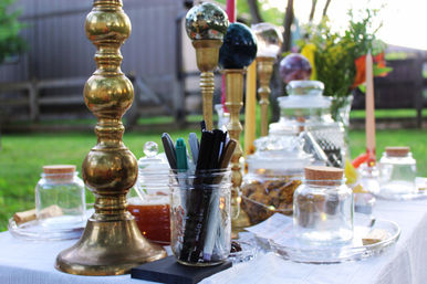 Backyard table display with tall brass candlesticks, decorative glass jars and a mason jar filled with pens and markers on a white cloth in soft evening light.