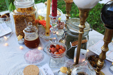 Outdoor boho tabletop display in a garden: glass jars of dried herbs and honey, brass candlesticks with lit taper candles, bowls of polished orange stones and rose quartz, tiny fairy lights on white linen.