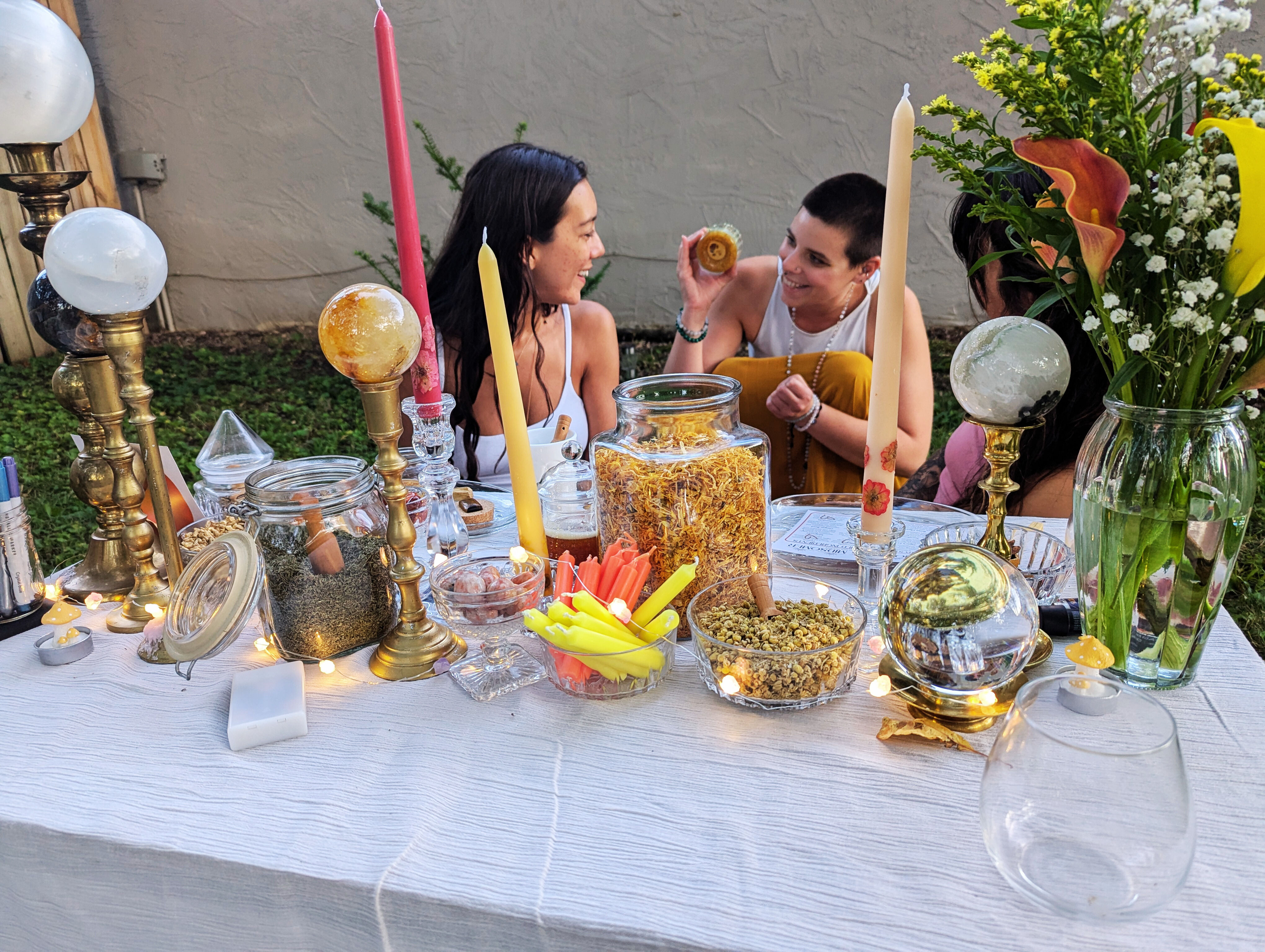 Backyard apothecary table with jars of dried herbs and flowers, brass candlesticks and crystal balls, colorful taper candles, a vase of blooms and fairy lights, three people chatting in the background.