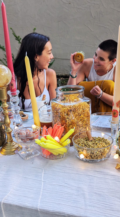 Backyard apothecary table with jars of dried herbs and flowers, brass candlesticks and crystal balls, colorful taper candles, a vase of blooms and fairy lights, three people chatting in the background.