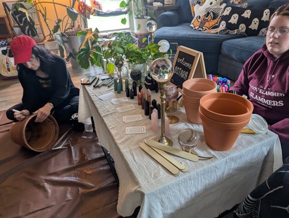 Cozy living-room DIY potting workshop with terracotta pots, crystals and glass spheres on a low table, plant cuttings, markers and wooden labels while two people prep a clay pot on the floor.