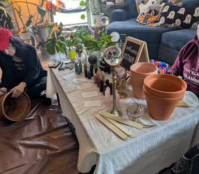 Cozy living-room DIY potting workshop with terracotta pots, crystals and glass spheres on a low table, plant cuttings, markers and wooden labels while two people prep a clay pot on the floor.