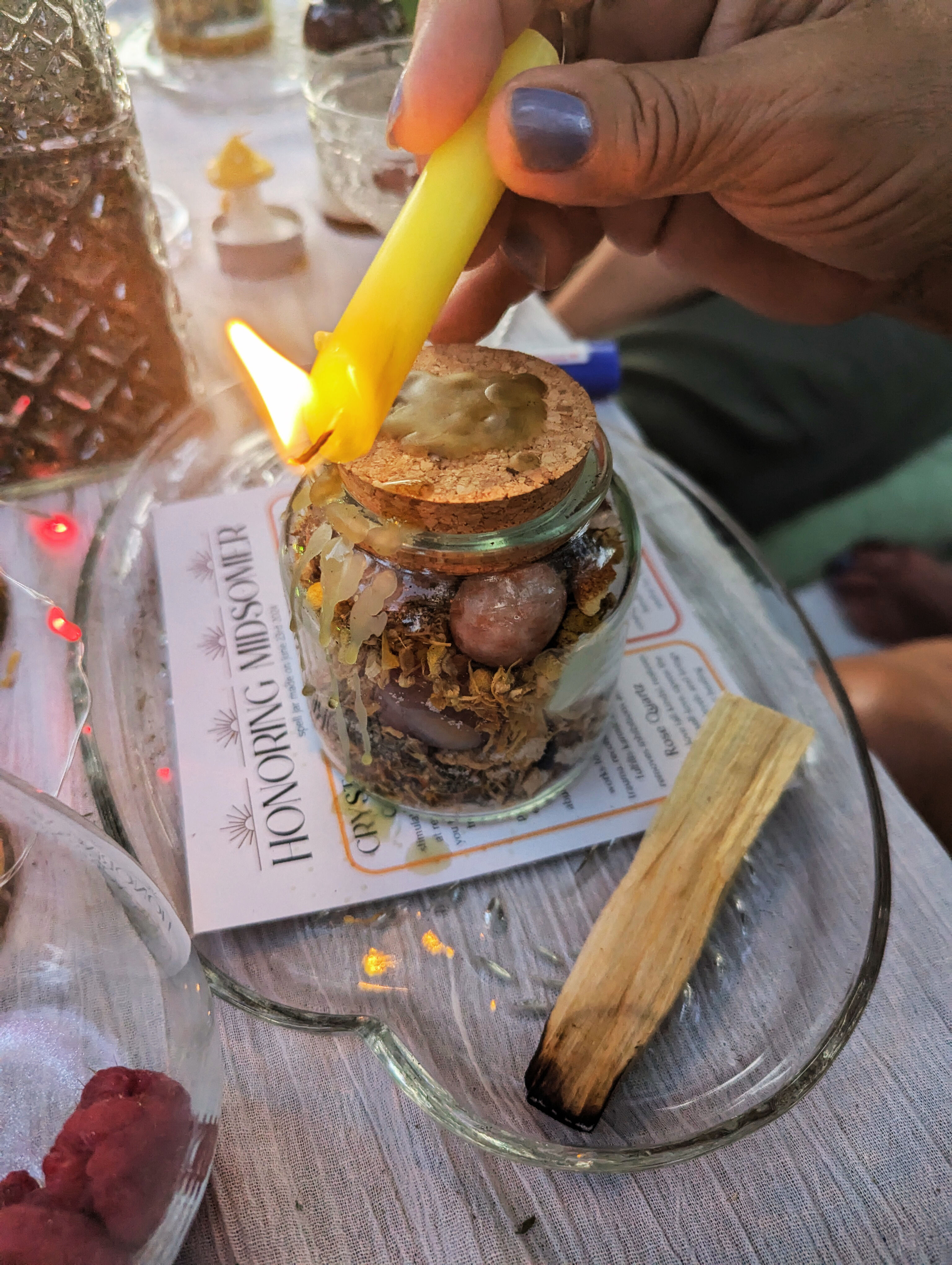 Hand holding a lit yellow candle to drip wax onto a cork-topped jar filled with dried herbs and crystals, with a charred palo santo stick on a glass tray nearby.