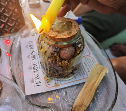 Hand holding a lit yellow candle to drip wax onto a cork-topped jar filled with dried herbs and crystals, with a charred palo santo stick on a glass tray nearby.