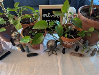 Cheerful indoor plant display: pothos vines in terracotta pots arranged with crystal towers, decorative orbs and a small "welcome" chalkboard on a white tablecloth.