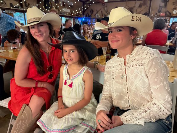 Smiling trio at a lively country-style saloon — two women and a young girl wearing cowboy hats; one in a red dress and boots, the girl in a white embroidered sundress, and the other in a white lace blouse holding a bottle, with string lights and a crowded wooden interior behind them.