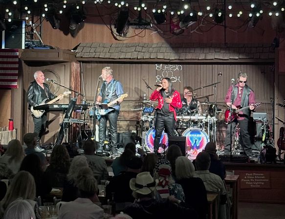Five-piece rock band performing on a rustic indoor stage under string lights — lead singer in a red jacket, two guitarists, keyboardist and drummer with colorful eagle bass drums, while a seated crowd watches and an American flag hangs to the side.