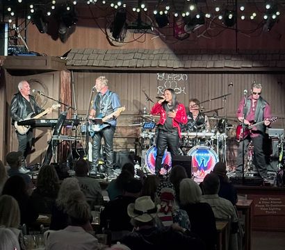 Five-piece rock band performing on a rustic indoor stage under string lights — lead singer in a red jacket, two guitarists, keyboardist and drummer with colorful eagle bass drums, while a seated crowd watches and an American flag hangs to the side.
