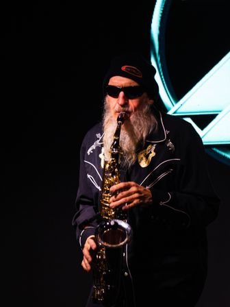 Bearded saxophonist in sunglasses and a beanie playing a shiny saxophone on a dark stage with teal geometric light backdrop