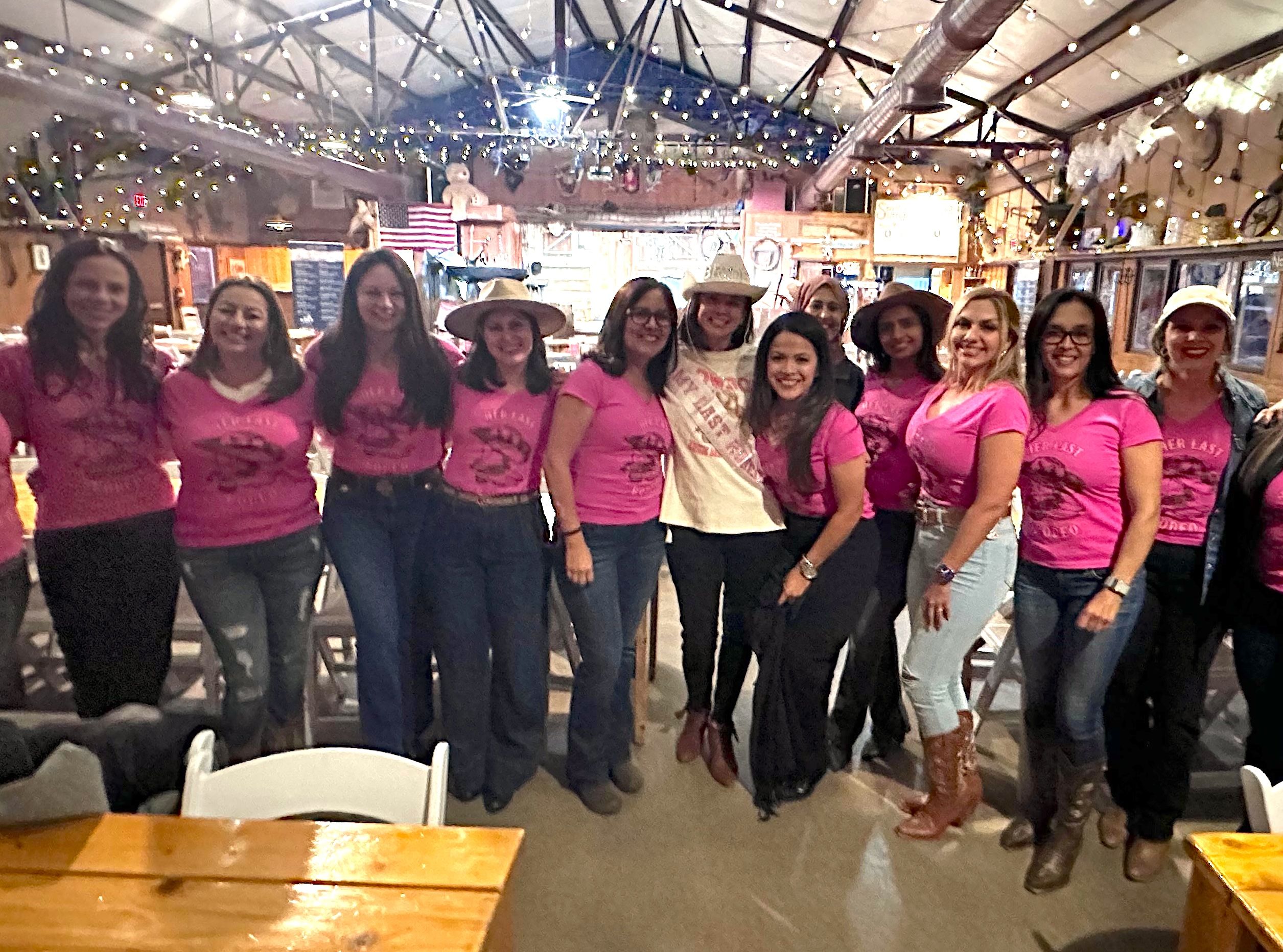 Group of women in matching pink shirts and cowboy hats posing with a bachelorette wearing a sash and straw hat inside a rustic barn-style venue with string lights, wooden tables and an American flag in the background.