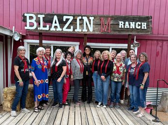 Smiling group of adults wearing red scarves posing on a wooden deck in front of a red barn-style ranch building and rustic sign, with hay bales, string lights and one person in a decorative jacket.