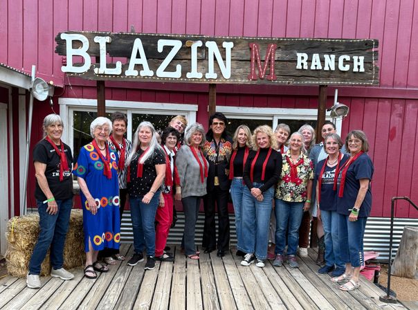 Smiling group of adults wearing red scarves posing on a wooden deck in front of a red barn-style ranch building and rustic sign, with hay bales, string lights and one person in a decorative jacket.