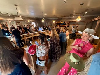 Busy western-style boutique interior with shoppers gathered around wooden counters trying on cowboy and cowgirl hats and accessories under warm pendant lights — lively hat-fitting event with rustic decor.