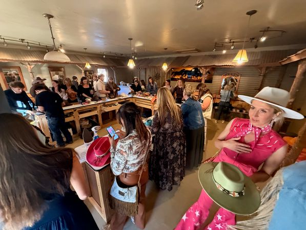 Busy western-style boutique interior with shoppers gathered around wooden counters trying on cowboy and cowgirl hats and accessories under warm pendant lights — lively hat-fitting event with rustic decor.