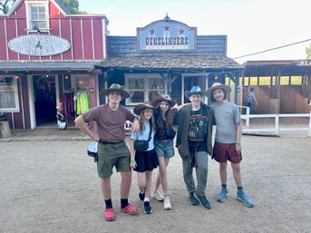 Five teens in cowboy hats pose on a dirt street in front of rustic Old West storefronts at a western-themed attraction.