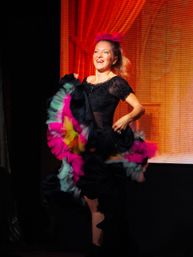 Smiling stage performer in black dress lifts a colorful ruffled skirt in a can-can style on a theater stage with a warm red curtain backdrop.