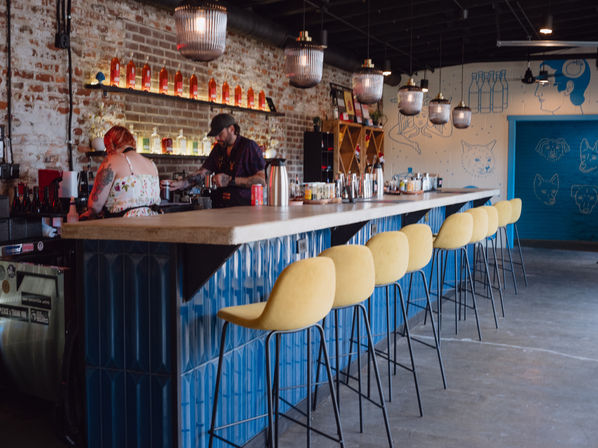 Interior of an urban cocktail bar with exposed brick wall, blue-tiled bar front and concrete countertop, a row of yellow bar stools, hanging pendant lights, back shelves of colorful bottles and two bartenders prepping drinks.
