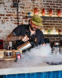 Tattooed bartender in a green cap pours liquid to create billowing fog for a dramatic dry-ice cocktail at an exposed brick bar with bottles on shelves.