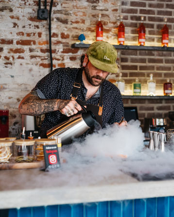 Tattooed bartender in a green cap pours liquid to create billowing fog for a dramatic dry-ice cocktail at an exposed brick bar with bottles on shelves.
