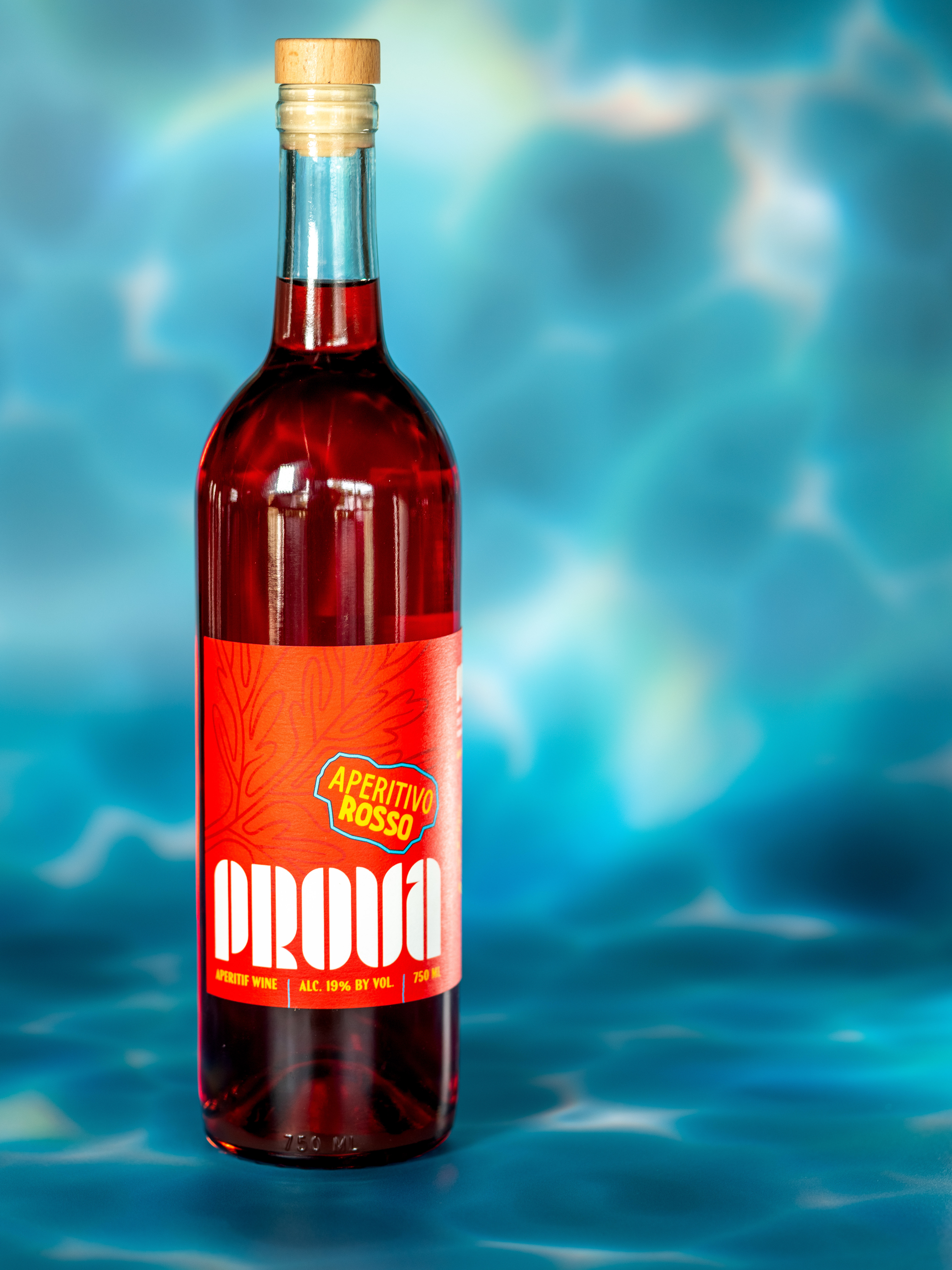 Ruby-red aperitif wine bottle with wooden cork and bright red label, staged on a turquoise pool-like background for a summery cocktail vibe.