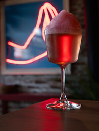 Frothy pink cocktail in a stemmed glass on a wooden bar top in a neon-lit bar interior, red neon sign and exposed brick wall blurred in the background.
