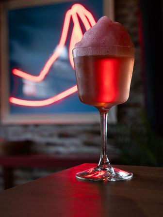 Frothy pink cocktail in a stemmed glass on a wooden bar top in a neon-lit bar interior, red neon sign and exposed brick wall blurred in the background.
