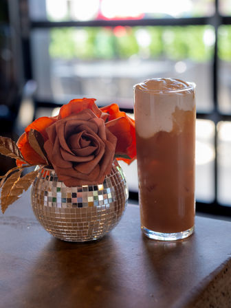 Tall creamy iced coffee in a clear glass next to a mirrored disco-ball vase holding orange-brown artificial roses on a cafe table with a blurred window background.