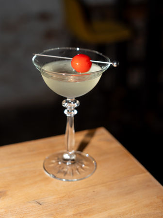Pale craft cocktail in an elegant coupe glass topped with a bright red cherry on a metal pick, sitting on a wooden table with a dark bar background.